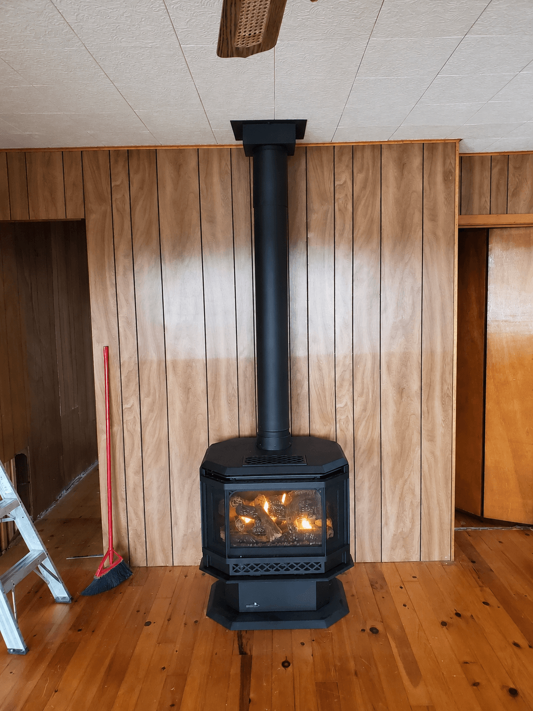 Black gas fireplace with glowing logs against a wood-paneled wall on a hardwood floor.