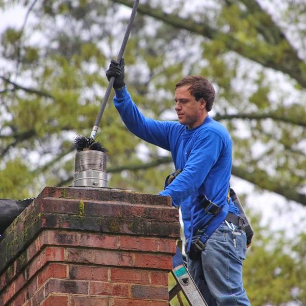 Man in blue shirt cleans a brick chimney using a long brush and safety harness.