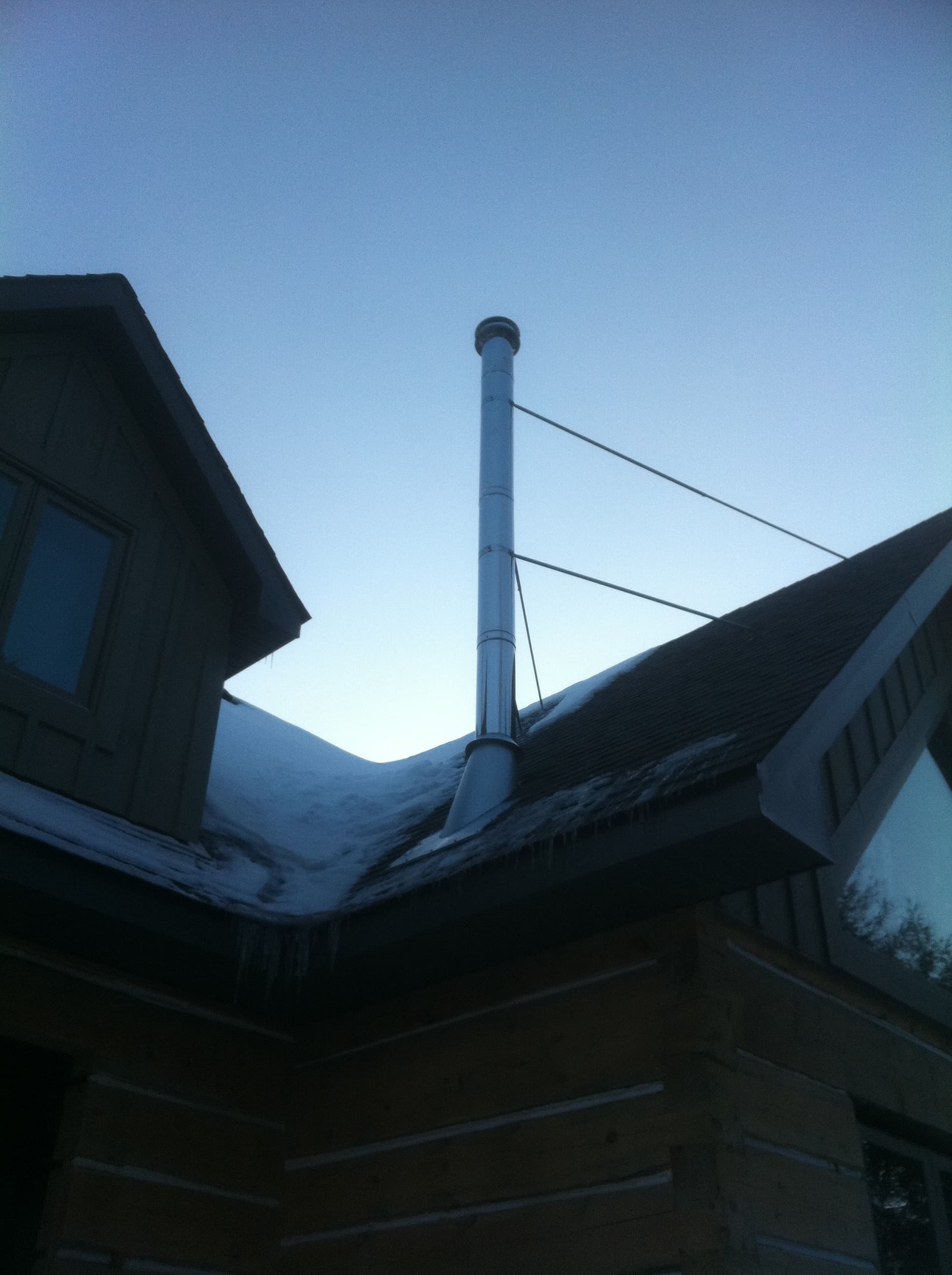 Tall silver chimney pipe with support wires on a snow-covered roof against a clear sky.