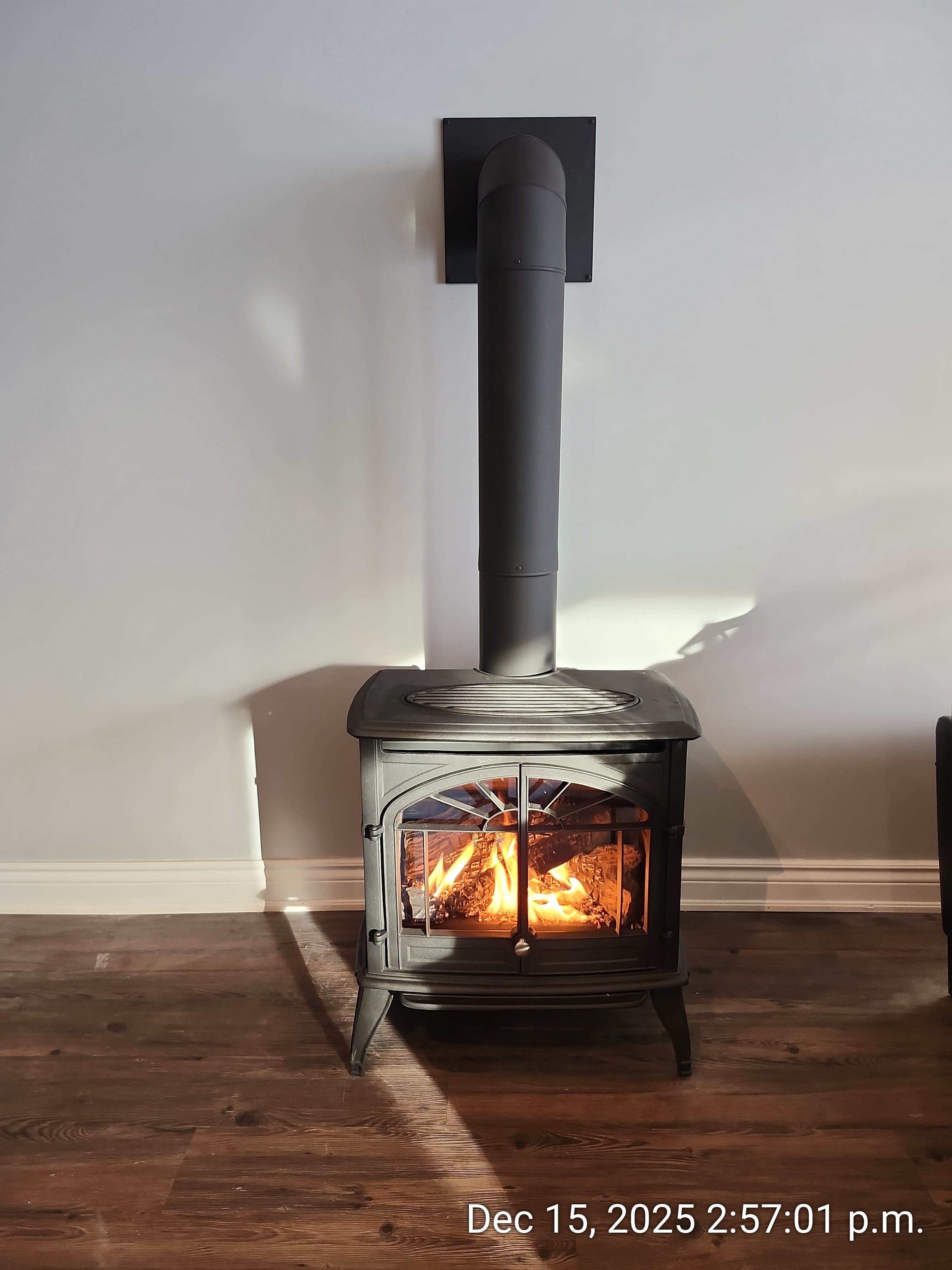 Black cast-iron stove with glowing orange flames inside, a tall flue pipe, and dark wood floors.