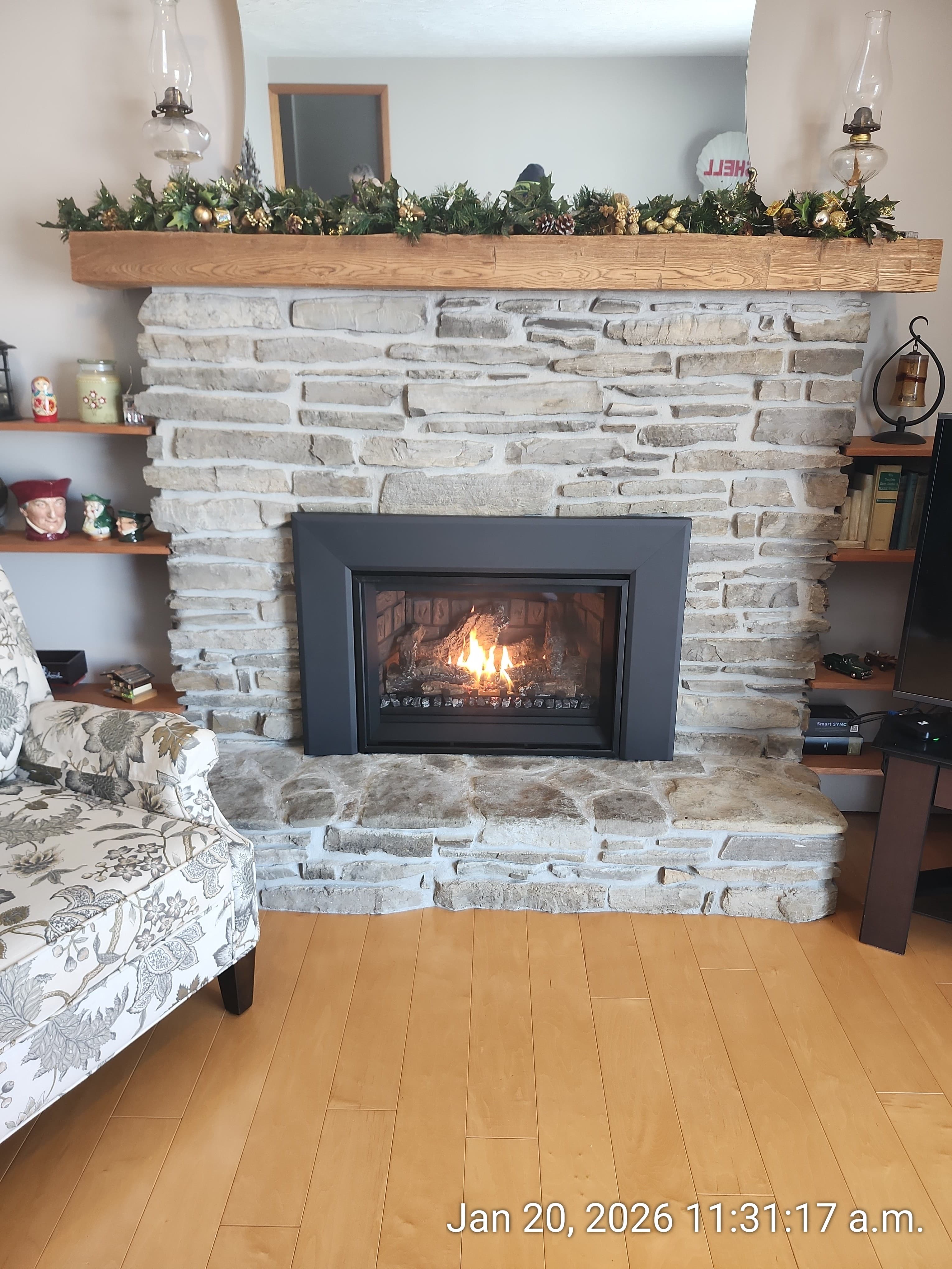 Stone fireplace with a burning fire, decorated wooden mantle, and floral armchair on wood floors.
