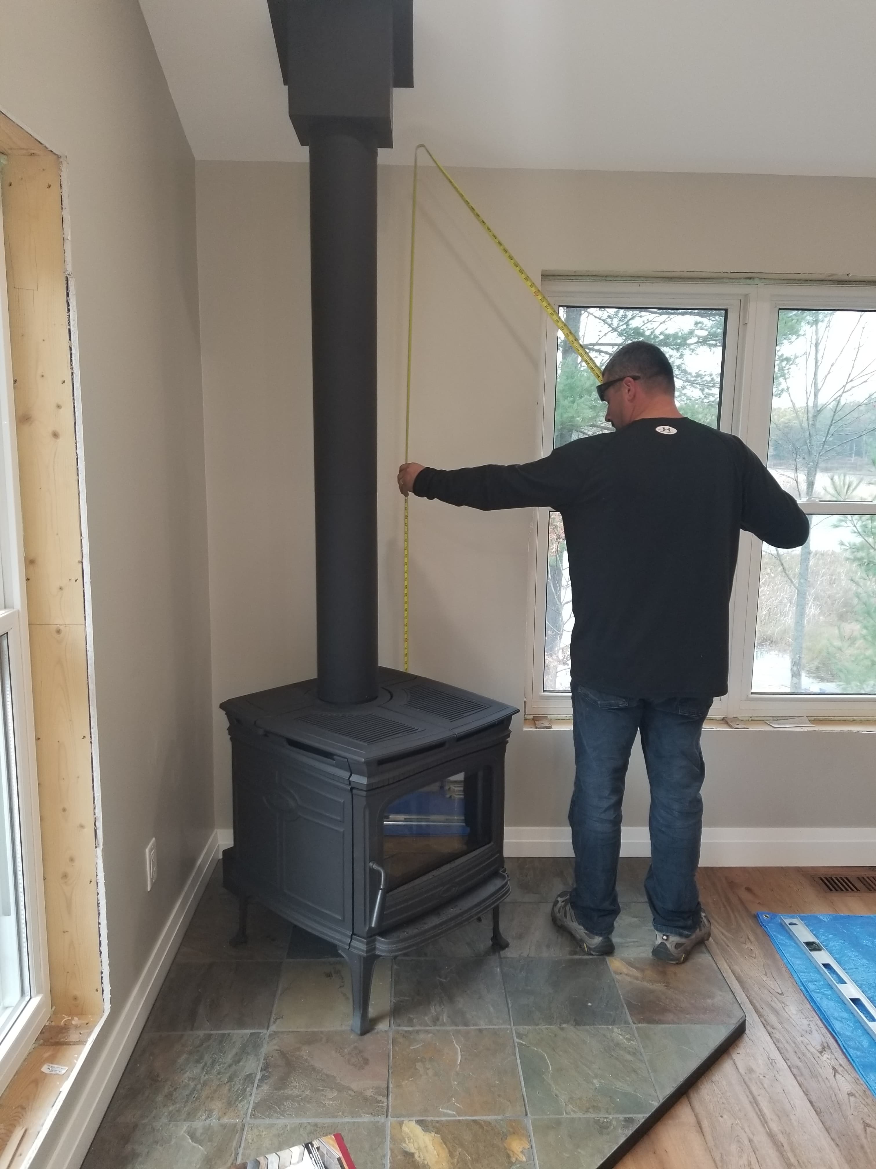 Man measuring distance between black wood stove pipe and window with yellow tape measure.