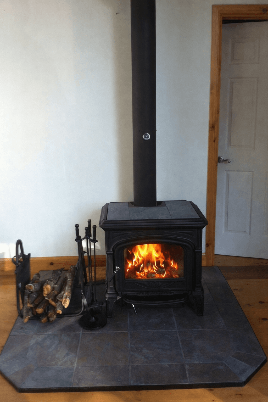 Black cast iron wood stove with a bright fire, next to logs and fireplace tools.