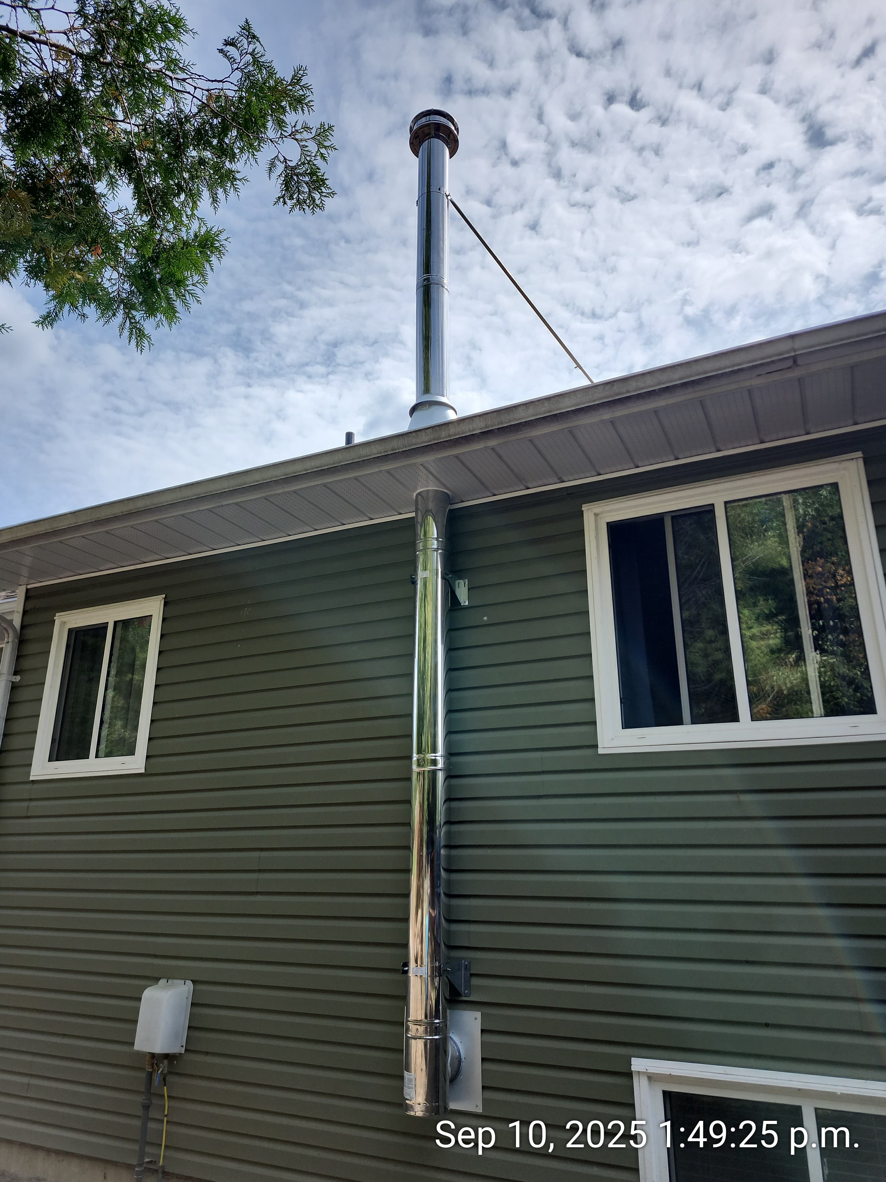 Tall stainless steel chimney pipe attached to green siding between two white-framed windows.