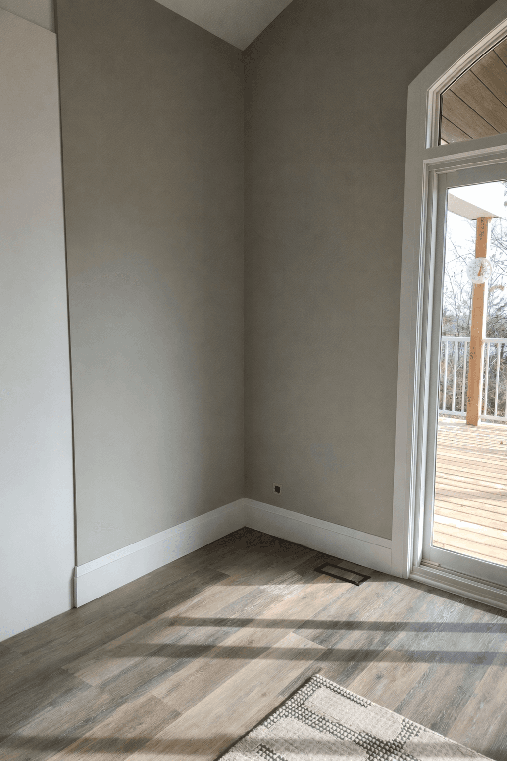 Empty room corner with greige walls, white baseboards, and sunlight streaming across wood floors.