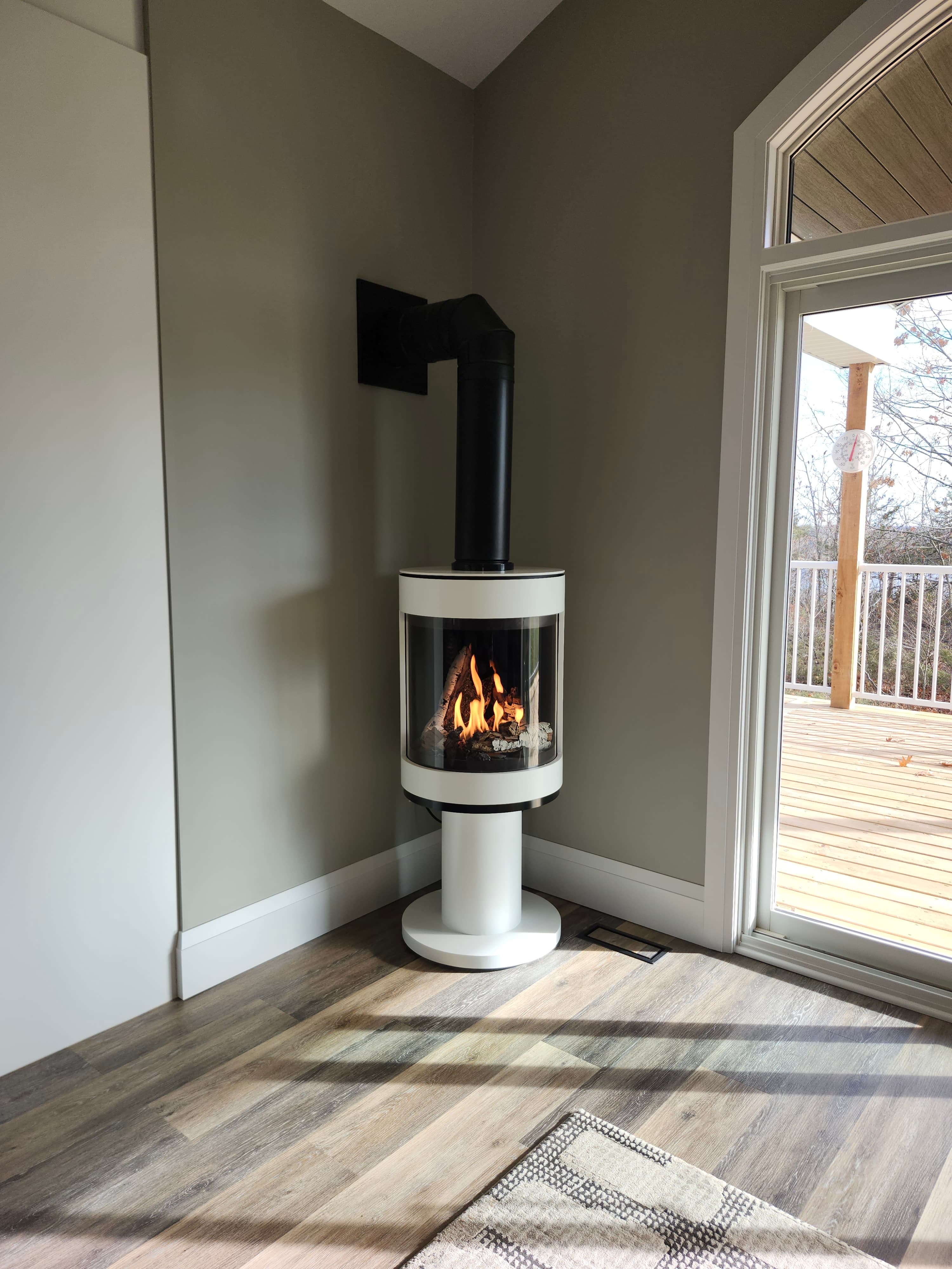 Modern white cylindrical wood stove with burning logs, positioned in a bright, sunlit room.