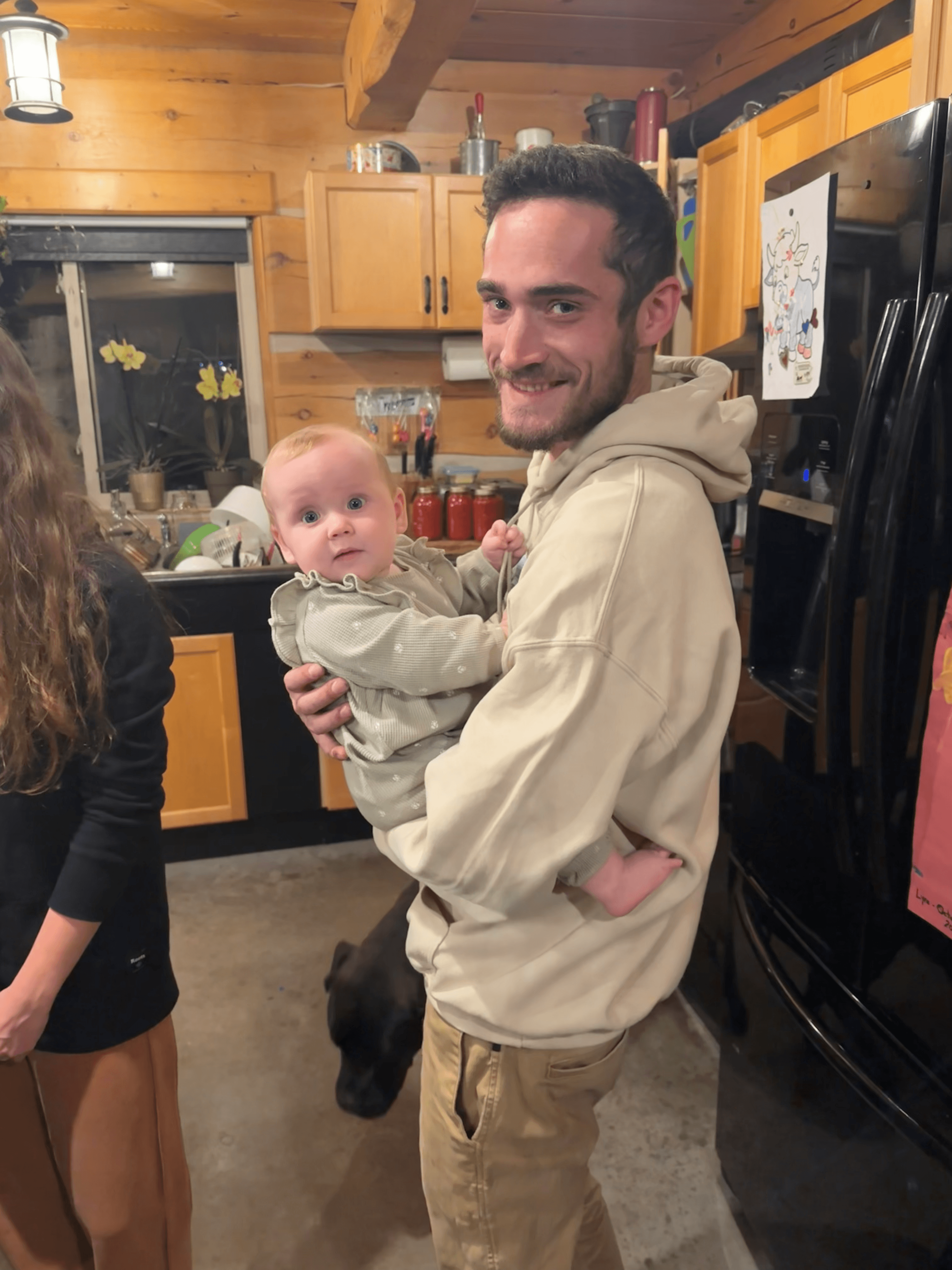 Smiling man in beige hoodie holds a baby in a green outfit in a kitchen.
