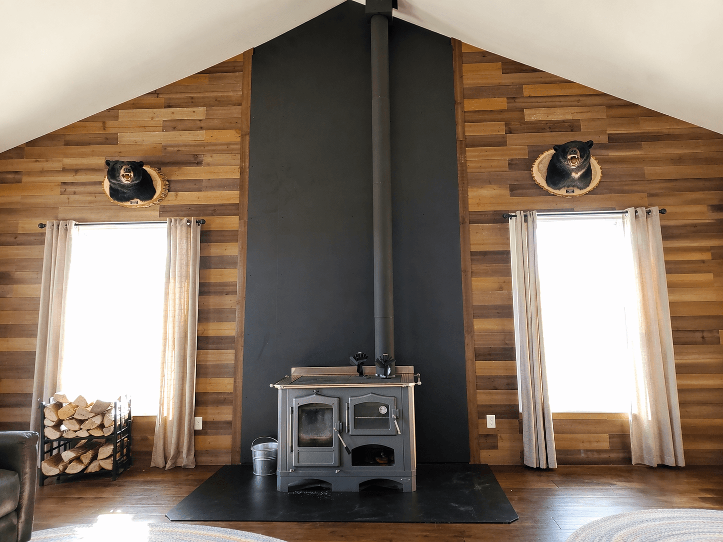 Wood stove centered between windows with mounted bear heads on a rustic, wood-paneled wall.