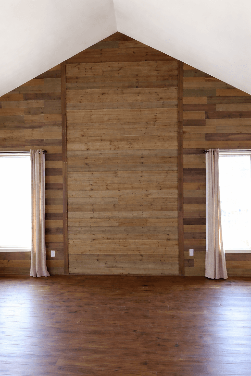 Empty room featuring a peaked wooden plank accent wall, vaulted ceiling, and two curtained windows.