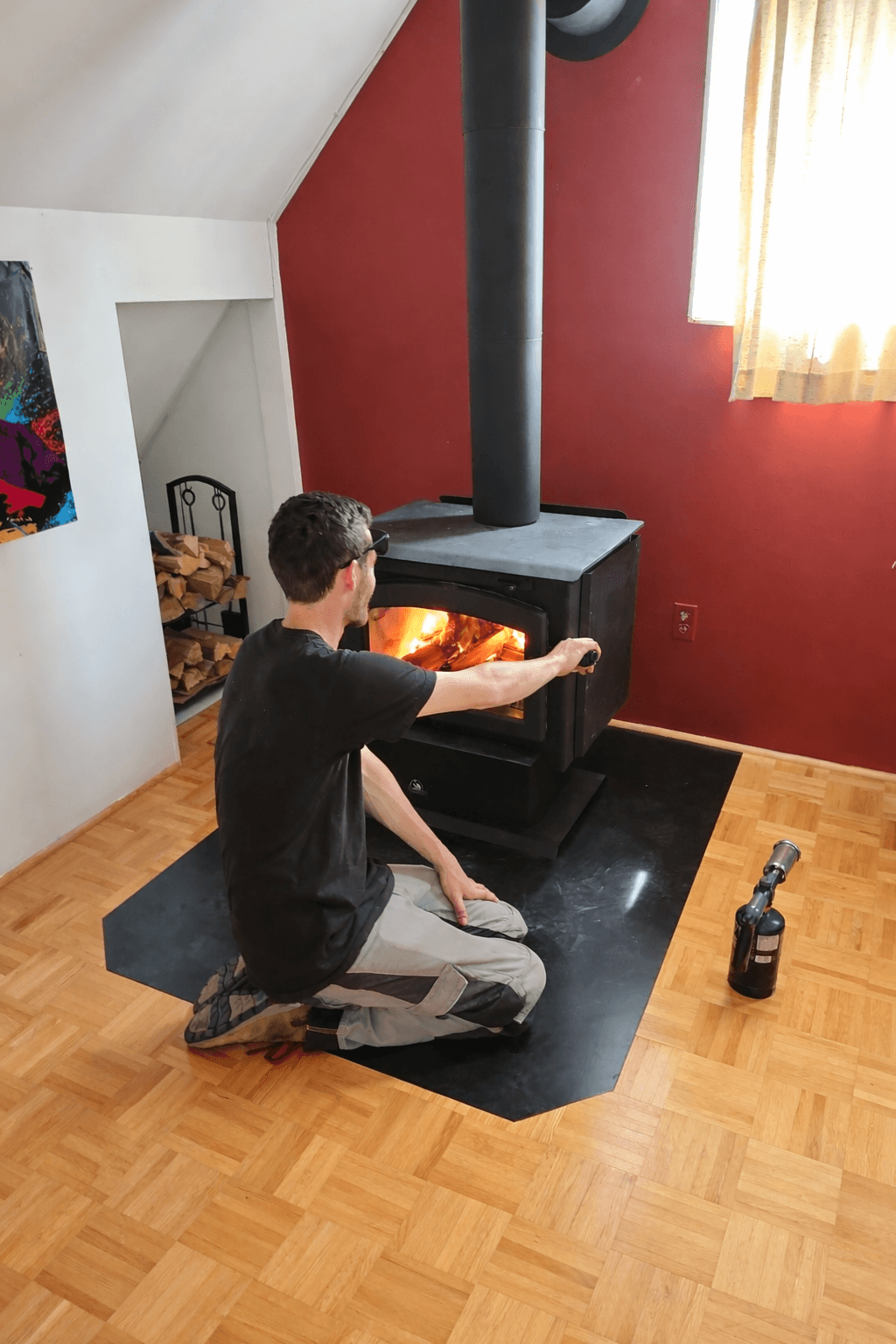 Man kneeling on a black mat, reaching to open a wood-burning stove with a glowing fire.
