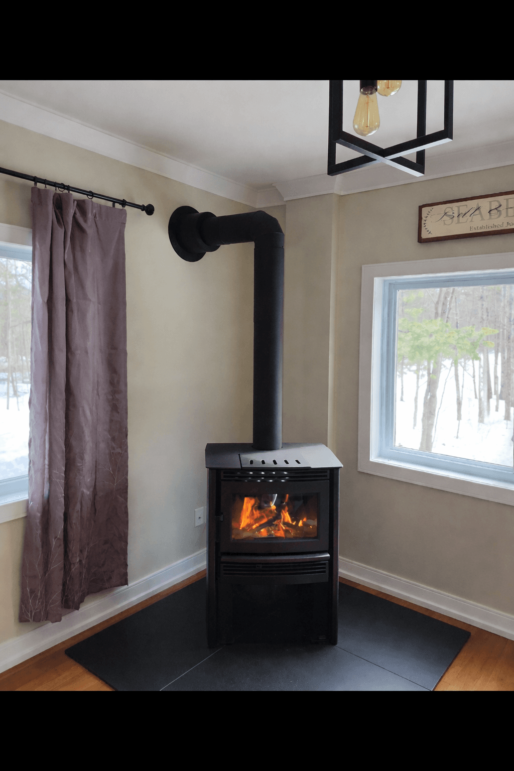 Black wood-burning stove with a glowing fire, positioned in a room corner between two bright windows.